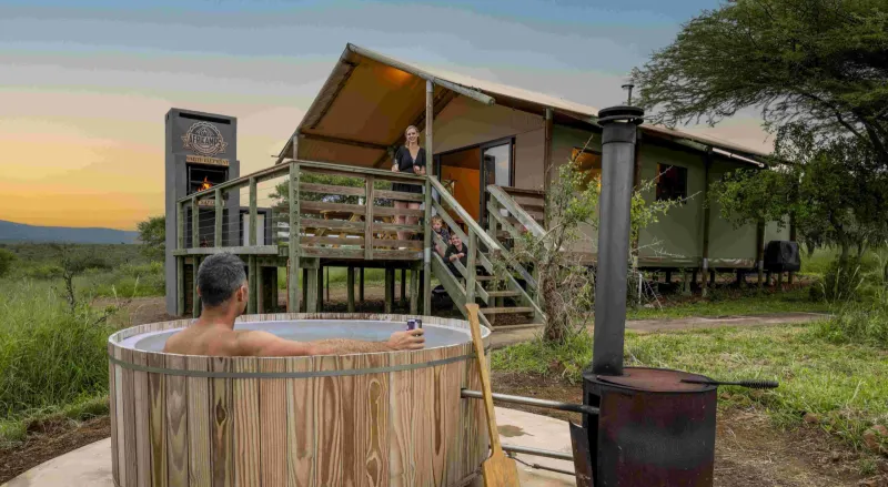 Man soaking in wooden hot tub outside safari tent lodge at sunset