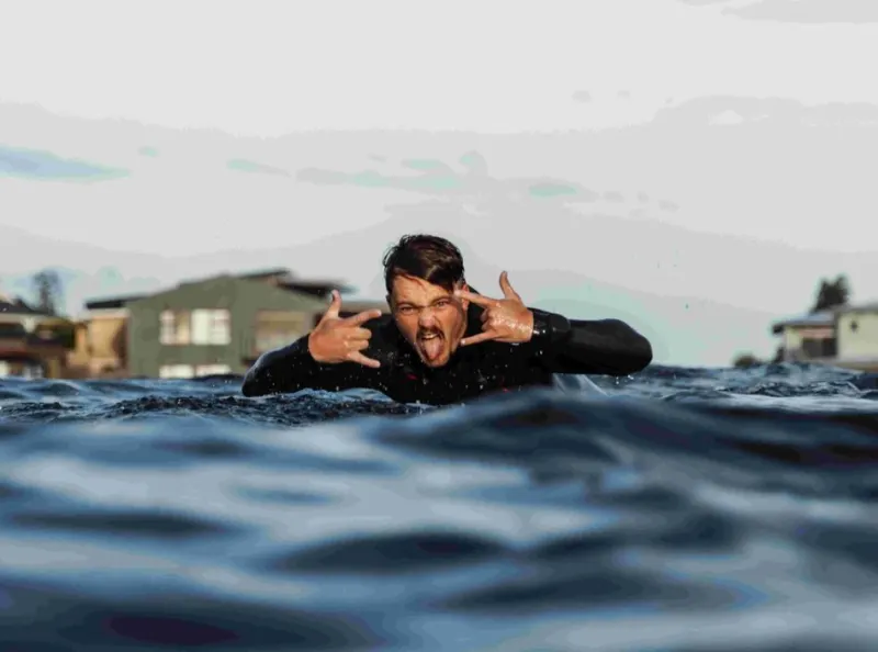 man in wetsuit lying on surfboard making hand gestures and sticking out tongue